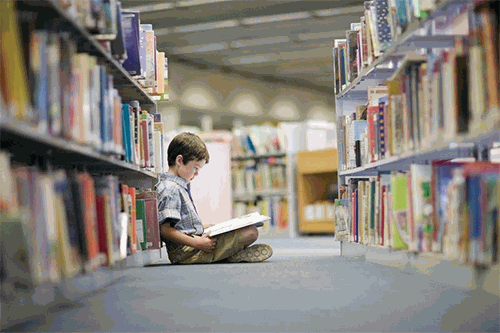 Photo of boy sitting down on carpet in library aisle reading a book